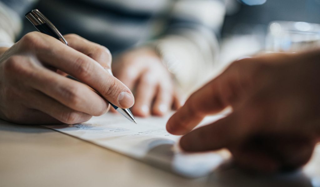Close up of unrecognizable man signing a contract while financial advisor is aiming at the place he need to sign.